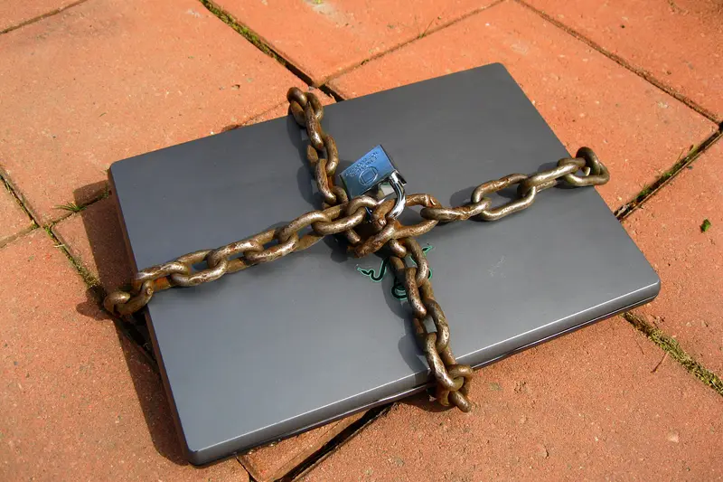 A laptop computer wrapped in a chain and secured with a padlock, photographed outdoors on brick paving